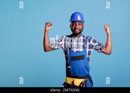 Operaio di costruzione afroamericano orgoglioso dei muscoli sviluppati del braccio dal lavoro pesante che esegue quotidiano. Il costruttore indossa tute e elmetto in studio girato su sfondo blu. Foto Stock