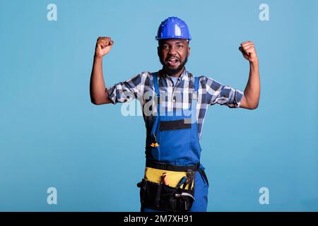 Operatore edilizio afro-americano in tute che mostra i muscoli del braccio davanti alla telecamera. Lavoratore di costruzione in elmetto dimostrando la sua forza contro sfondo blu in studio shot. Foto Stock