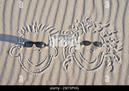 Faccia disegnata nella sabbia sulla spiaggia, con gli occhiali da sole. Sabbia con modello di onda. Foto vacanza primordiale dal mare Foto Stock