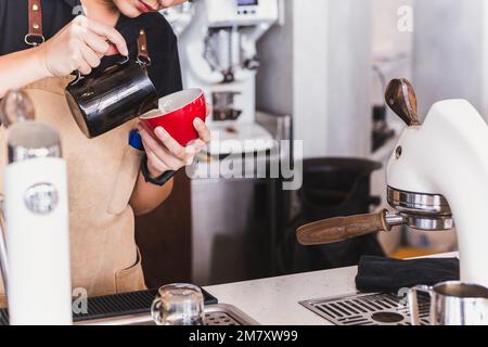 Primo piano della barista asiatica che prepara il cappuccino facendo latte art in tazza. Foto Stock