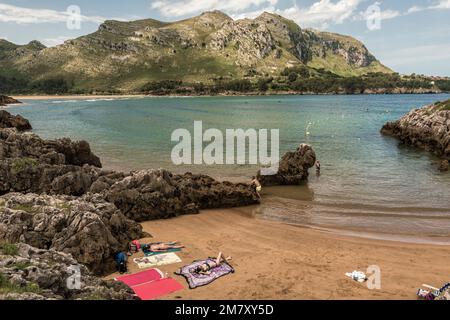 Due donne stese sull'asciugamano in bikini sulla sabbia della piccola spiaggia della città di Islares, mentre altri bagnano, Cantabria, Spagna Foto Stock