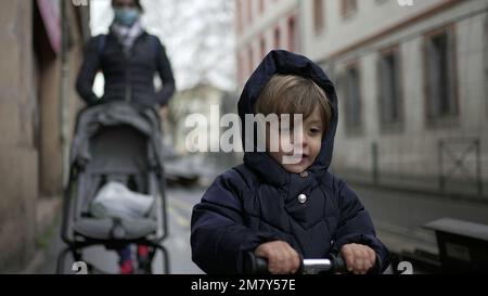 Piccolo bambino in sella a uno scooter a tre ruote sul marciapiede urbano della città con la madre che cammina Foto Stock