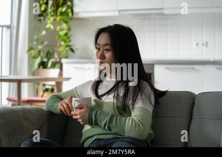 Giovane preoccupata donna asiatica penosa seduta sul divano a casa con una tazza di tè preoccupata di qualcosa Foto Stock