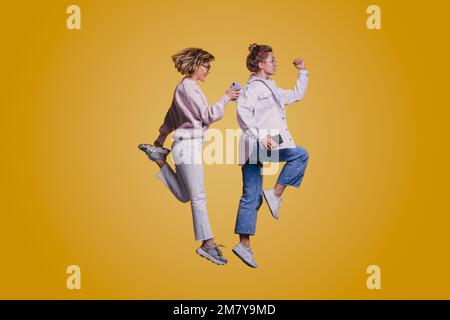 Vista a tutta lunghezza delle dimensioni del corpo di due donne allegre che saltano tenendo i loro telefoni isolati su sfondo di colore giallo Foto Stock