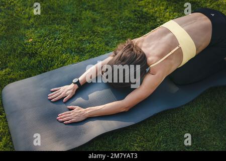 Vista dall'alto di una donna sportiva che le stracciava la schiena prima di altri exersices consigliati, seduto sul tappetino yoga all'aria aperta. Effetto riflesso del sole. Stile di vita felice Foto Stock