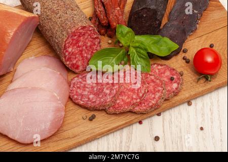 Diversi tipi di salsicce prosciutto, mahan, salame su un asse di legno decorato con pomodori e basilico Foto Stock