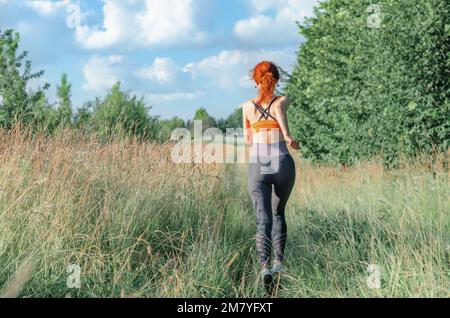 Una giovane runner femminile corre lungo la strada coltivata con erba. Vista posteriore Foto Stock