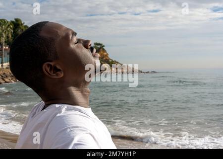 Vista laterale di afro americano maschio con gli occhi chiusi godendo aria fresca vicino al mare sulla spiaggia durante le vacanze estive Foto Stock