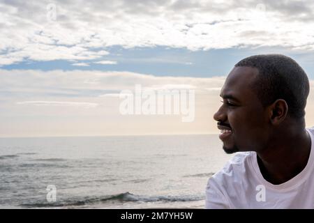 Vista laterale del giovane afro-americano maschio in t shirt bianca sorridente e guardando lontano vicino mare ondeggiante sulla spiaggia contro cielo nuvoloso Foto Stock