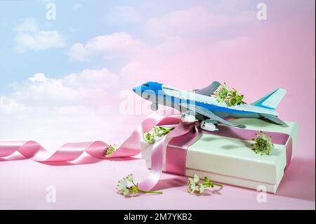 Aeroplano giocattolo su una scatola regalo con nastro e alcuni fiori, sfondo rosa che svanisce in un cielo blu nuvoloso, viaggio e viaggio presente per San Valentino Foto Stock