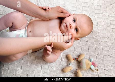 La madre massaggia e lubrifica delicatamente il viso del bambino. Mamma che applica crema o olio sul viso del bambino. Felice neonato sorridente. Neonato al Cozy H Foto Stock