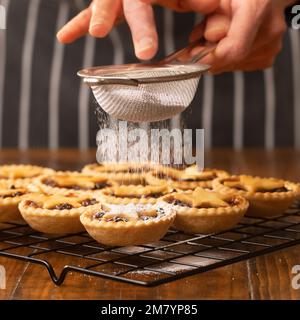 Torte tritate sormontate da stelle di pasta su una griglia metallica che viene dragata in zucchero a velo da una persona non identificabile in un grembiule a strisce nere. Foto Stock