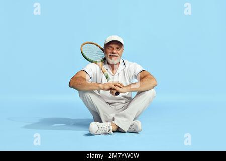 Delizioso. Ritratto di bell'uomo anziano in elegante vestito bianco seduto, in posa con racchetta da tennis su sfondo blu. Concetto di tempo libero Foto Stock