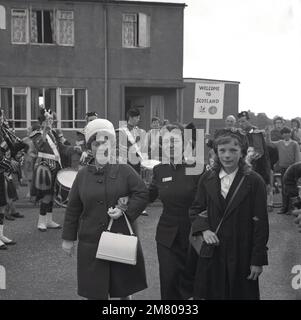 1960s, storica, band bagpipe suona per strada, come una signora del Salvation Army in Canada paga un vistor al twon/village, Fife, Scozia, UK. Foto Stock