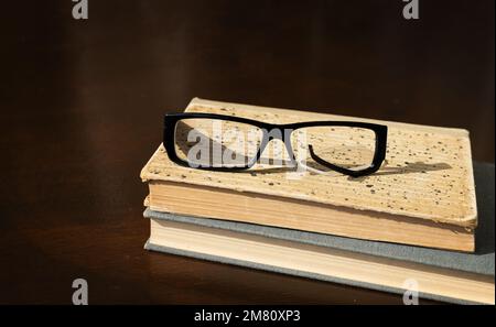 una cornice di occhiali rotta si trova su una pila di vecchi libri su un tavolo di legno marrone Foto Stock