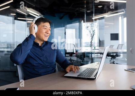 Felice giovane studente maschio, freelancer, blogger ascoltare musica in cuffie. Si siede in ufficio a un tavolo con un computer portatile. Pausa. riposi. Foto Stock