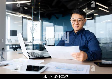 Un giovane uomo, un architetto asiatico, un designer, un ingegnere, un freelance lavora in ufficio alla scrivania utilizzando un computer portatile. Contiene documenti, piani e disegni nelle sue mani. Foto Stock