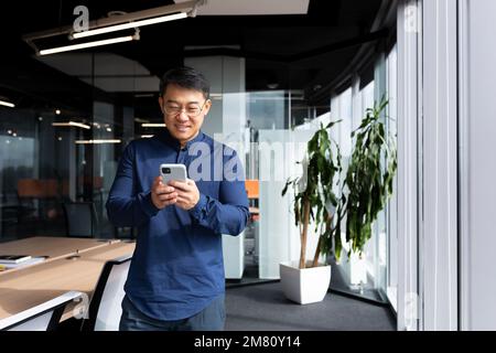 Un giovane uomo, un uomo d'affari asiatico, uno studente in piedi accanto alla finestra in ufficio, utilizza un telefono cellulare, digita messaggi, legge, controlla la posta. Guarda la telecamera, sorride. Foto Stock