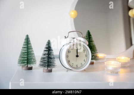 Una sveglia bianca, un puntatore a dodici, un piccolo albero di Natale artificiale verde e candele accese in candelabri su una cassettiera bianca. Copia Foto Stock