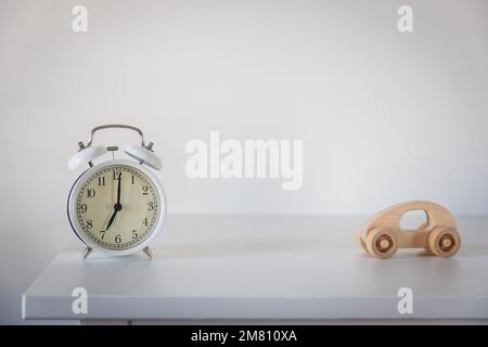 Sveglia bianca con frecce alle sette del mattino. Piccola auto in legno in stile scandinavo su una cassettiera bianca. Concetto - mattina, Foto Stock