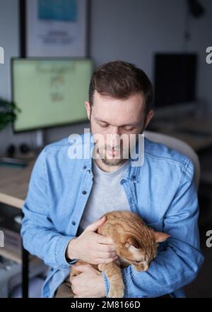Uomo caucasico freelance software engineer seduto al posto di lavoro in casa ufficio tenendo il gatto zenzero. Giovane uomo sviluppatore grigio in t-shirt con animale domestico. Immagini di alta qualità Foto Stock