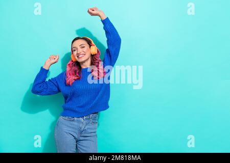 Foto di una ragazza soddisfatta con le cuffie maglione blu ricci dai capelli ricci che alzano i pugni danzando isolati su uno sfondo dai colori vivaci del teal Foto Stock