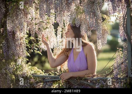 Donna matura felice premurosa circondata da glicine cinese in abito viola Foto Stock