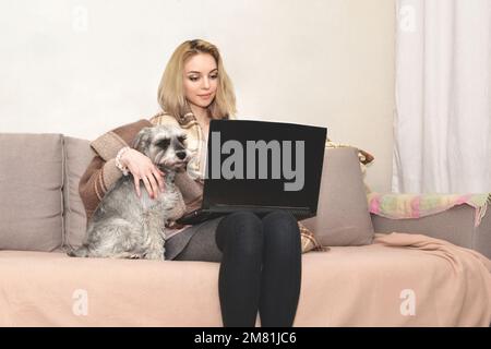 Una bella donna bionda è seduta a casa con un computer portatile sul divano e abbraccia un cane grigio carino. Foto Stock