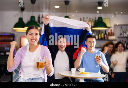 Compagnia di tifosi gioiosi che sventolano la bandiera della Russia e celebrano la vittoria della squadra di calcio nello sport bar Foto Stock