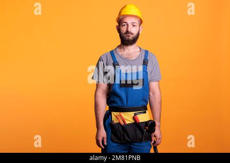 Sicuro lavoratore di costruzione indossando uniforme rinnovante e hardHat, posando sulla macchina fotografica in studio. Riparatore professionista che si prepara a lavorare su progetto di ristrutturazione, artigiano esperto. Foto Stock