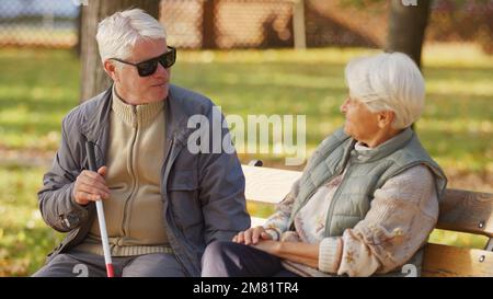 Uomo anziano cieco con occhiali da sole a riposo con la moglie su una panchina nel parco. Foto di alta qualità Foto Stock