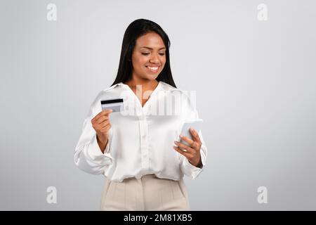 Felice donna d'affari nera millennial in blusa bianca legge il messaggio sul telefono usa la carta di credito Foto Stock