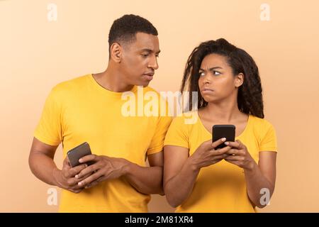 Ragazzo afro-americano con smartphone spionaggio su sua moglie chattando sul telefono cellulare, sfondo giallo studio Foto Stock