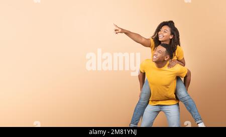 Eccitato coppia nera che ha divertimento, ragazzo piggybacking signora e indicando lo spazio di copia, sfondo giallo, panorama Foto Stock