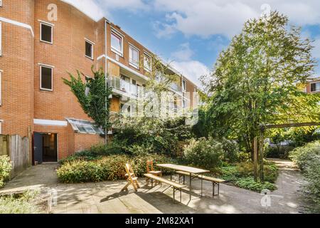 un'area esterna con tavoli, sedie e alberi di fronte ad un edificio di appartamenti in mattoni in una giornata di sole cielo blu Foto Stock