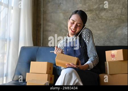 Affascinante e occupato imprenditore asiatico di affari di e-commerce femminile o venditore online è al telefono con i suoi fornitori, mentre la preparazione shippin Foto Stock