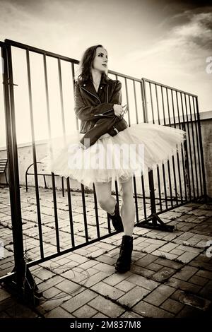 una ballerina in tutu si trova al recinto sul tetto al tramonto in stivali e una giacca Foto Stock