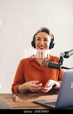 Felice podcaster femmina sorridente mentre si siede dietro un microfono e un computer portatile con cuffie. Donna allegra che ospita un podcast su Internet nella sua casa Foto Stock