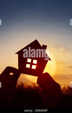 Mani che tengono la casa di carta sul cielo del tramonto, casa di famiglia, casa senza tetto, e concetto di assicurazione di protezione della casa, giorno internazionale delle famiglie, nuova vita Foto Stock