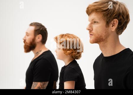 vista laterale del ragazzo rosso vicino a papà e nonno barbuto in magliette nere isolato su grigio, immagine stock Foto Stock