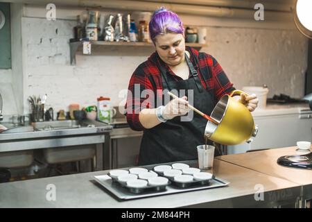 medium shot di un cuoco femmina con i capelli viola che mettono l'impasto da una ciotola in una tazza da aggiungere ai muffin al cioccolato. Foto di alta qualità Foto Stock