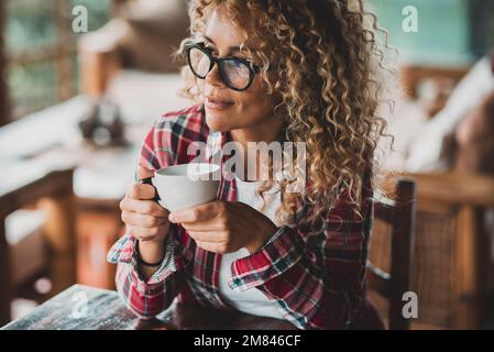 Attraente donna di mezza età con lunghi capelli ricci godendo di relax a casa bere tè o caffè da solo seduti al tavolo. Serena gente femminile portra Foto Stock