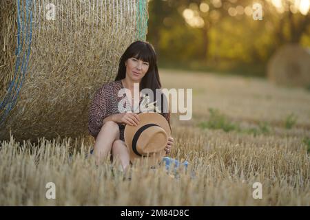 Una bella ragazza siede in un campo di fronte ad un pagliaio con un cappello nelle sue mani. Foto Stock