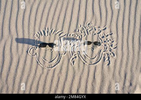 Faccia disegnata nella sabbia sulla spiaggia, con gli occhiali da sole. Sabbia con modello di onda. Foto vacanza primordiale dal mare Foto Stock