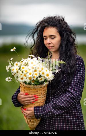 Una donna si trova su un campo verde e tiene in mano un cesto con un grande bouquet di margherite. Sullo sfondo sono montagne e un lago. Foto Stock