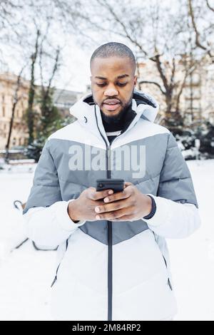 Giovane afroamericano in abiti casual bianco digitando messaggio, SMS al telefono, in parco o strada all'aperto. Stagione invernale con neve Foto Stock
