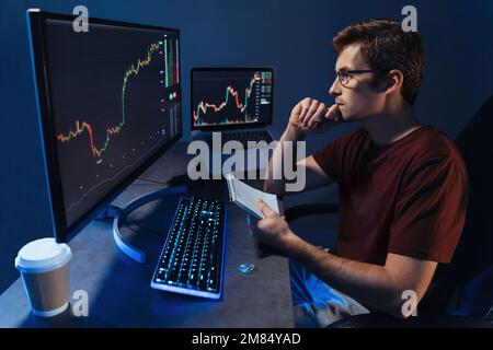 Crypto Investor lavora al pc, notando i risultati della sua analisi del mercato commerciale, seduto al tavolo, guardando la visualizzazione con concentrazione, analizzando i movimenti dei prezzi, adeguando la strategia di investimento Foto Stock