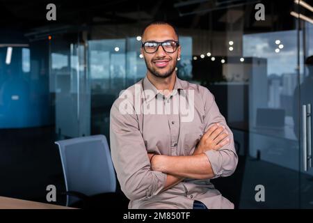 Ritratto di uomo d'affari ispanico riuscito, uomo riuscito in camicia che guarda la macchina fotografica con le braccia incrociate, operaio all'interno dell'ufficio vicino alla finestra. Foto Stock