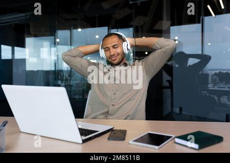 Giovane uomo d'affari afro-americano di successo che si rilassa in ufficio, uomo in camicia e cuffie ascoltando musica online radio e podcast, seduto alla scrivania utilizzando un computer portatile al lavoro. Foto Stock