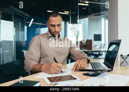 Serio uomo d'affari ispanico che lavora con documenti e fatture all'interno dell'ufficio, uomo dietro lavoro di carta, usando il laptop al lavoro seduto alla scrivania. Foto Stock